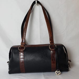 Puede incluir: Bolso de mano de cuero negro con detalles y asas marrones con textura de cocodrilo. El bolso presenta herrajes plateados, incluidos anillos y un dije en forma de coraz&oacute;n. El bolso tiene forma rectangular con dos asas.