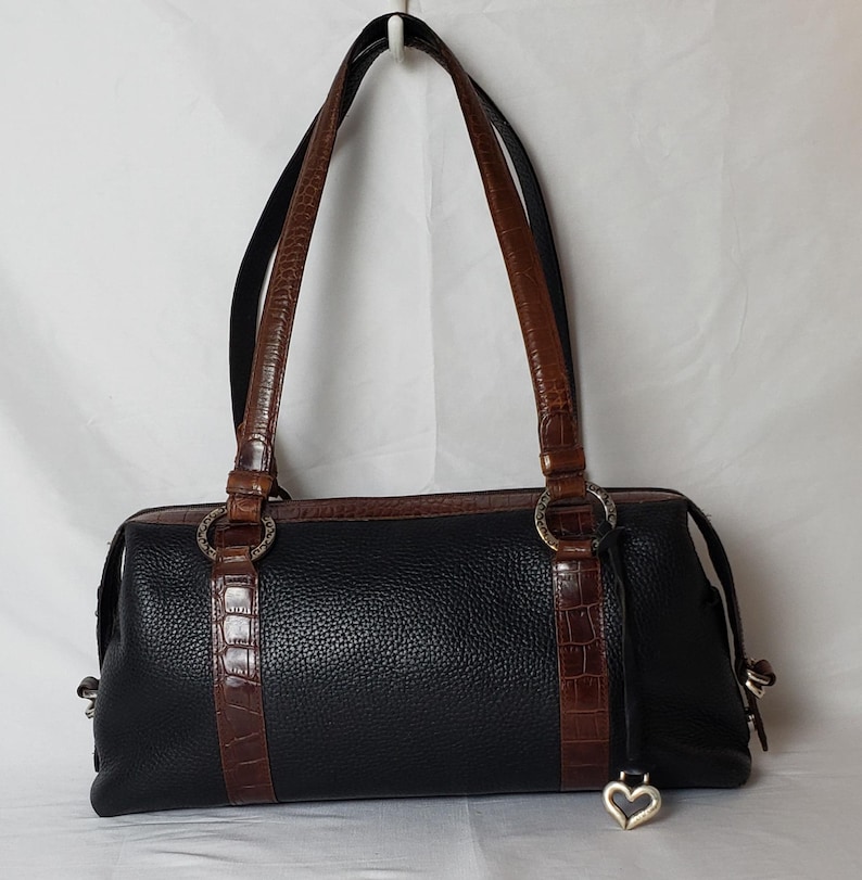 Puede incluir: Bolso de mano de cuero negro con detalles y asas marrones con textura de cocodrilo. El bolso presenta herrajes plateados, incluidos anillos y un dije en forma de coraz&oacute;n. El bolso tiene forma rectangular con dos asas.