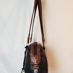 Puede incluir: Bolso de mano de cuero negro y marr&oacute;n con un acabado texturizado. El bolso presenta un detalle marr&oacute;n con relieve de cocodrilo en la parte superior y una hebilla a juego. Tiene dos correas de cuero marr&oacute;n y un charm plateado en forma de coraz&oacute;n.