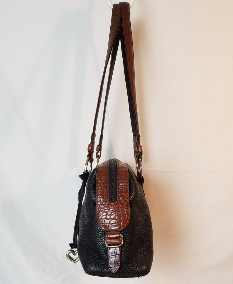 Puede incluir: Bolso de mano de cuero negro y marr&oacute;n con un acabado texturizado. El bolso presenta un detalle marr&oacute;n con relieve de cocodrilo en la parte superior y una hebilla a juego. Tiene dos correas de cuero marr&oacute;n y un charm plateado en forma de coraz&oacute;n.