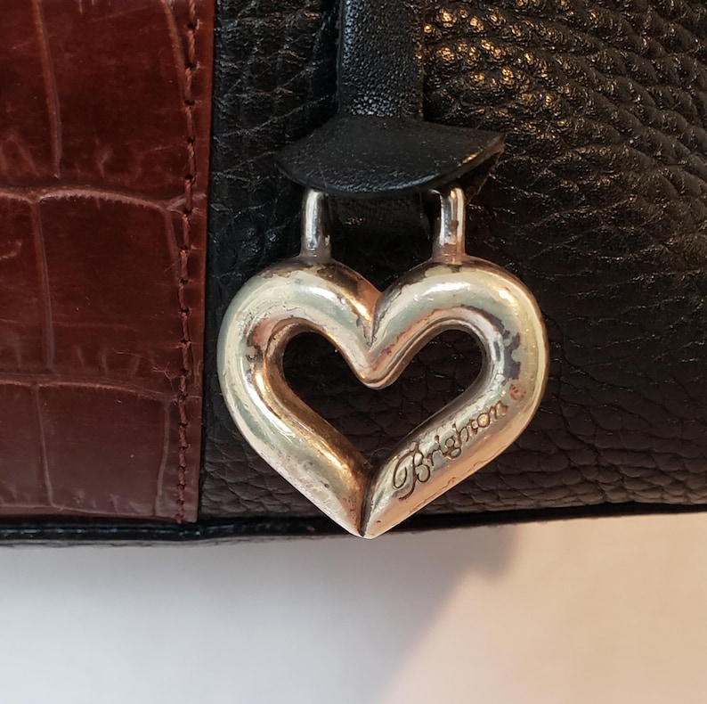Puede incluir: Primer plano de un bolso de mano negro, marr&oacute;n y plateado. El bolso presenta cuero negro texturizado, un panel marr&oacute;n con relieve de cocodrilo y un dije plateado en forma de coraz&oacute;n con la palabra "Brighton" grabada.