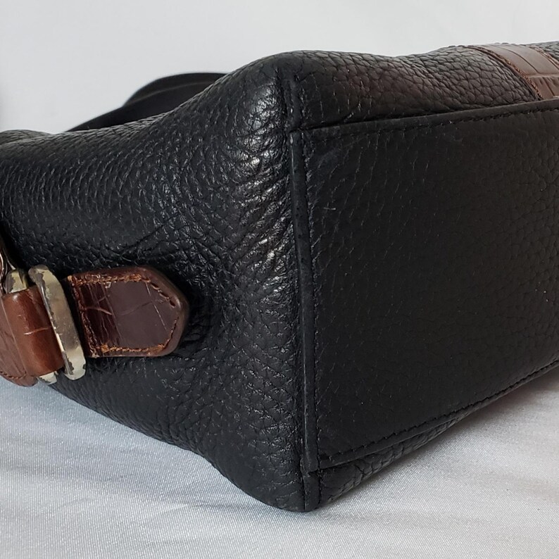 Puede incluir: Bolso de mano de cuero negro con acabado texturizado. Cuenta con detalles de cuero repujado con estampado de cocodrilo marr&oacute;n en la correa y la hebilla. El bolso tiene una hebilla plateada.