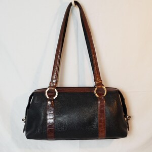 Puede incluir: Bolso de hombro de cuero negro con detalles y asas de cuero marr&oacute;n. El bolso presenta un exterior texturizado, una parte superior de cuero marr&oacute;n y anillos decorativos dorados. Las asas son largas y delgadas, y el bolso tiene forma rectangular.