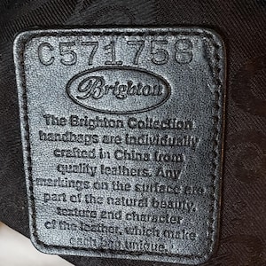 Puede incluir: Primer plano de una etiqueta de cuero negro con el logotipo de Brighton y el n&uacute;mero C571758. La etiqueta incluye el texto "The Brighton Collection handbags are individually crafted in China from quality leathers."