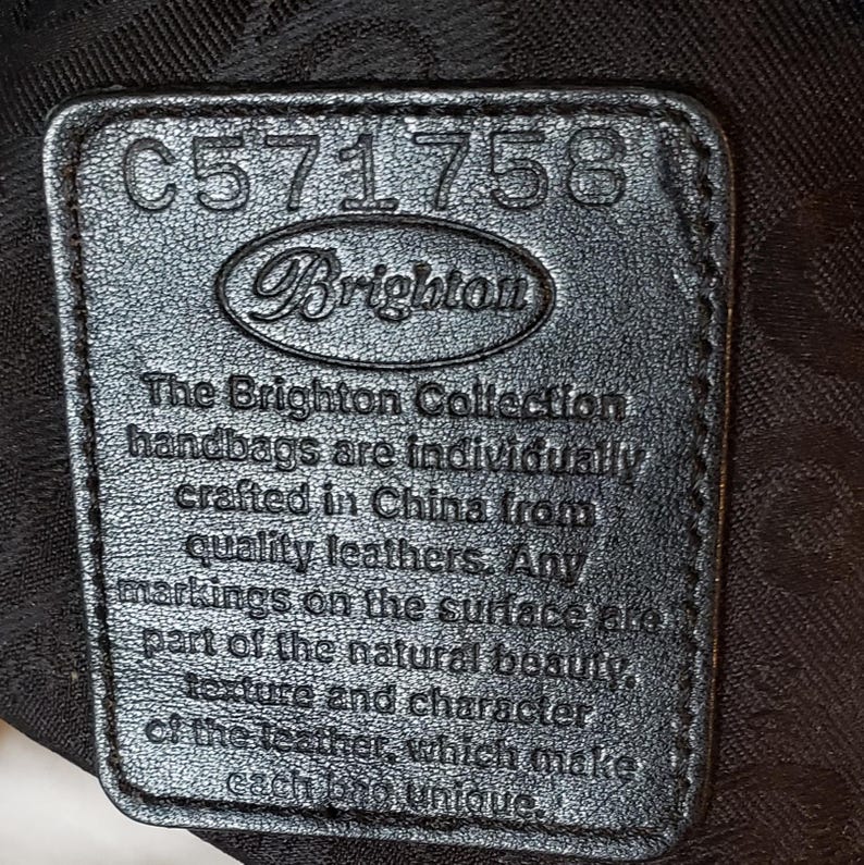 Puede incluir: Primer plano de una etiqueta de cuero negro con el logotipo de Brighton y el n&uacute;mero C571758. La etiqueta incluye el texto "The Brighton Collection handbags are individually crafted in China from quality leathers."