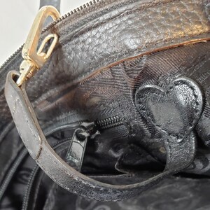 Puede incluir: Primer plano de un bolso de mano de cuero negro con un cierre y asa dorados. El bolso presenta un adorno en forma de coraz&oacute;n y un cierre de cremallera. El cuero tiene una apariencia texturizada.