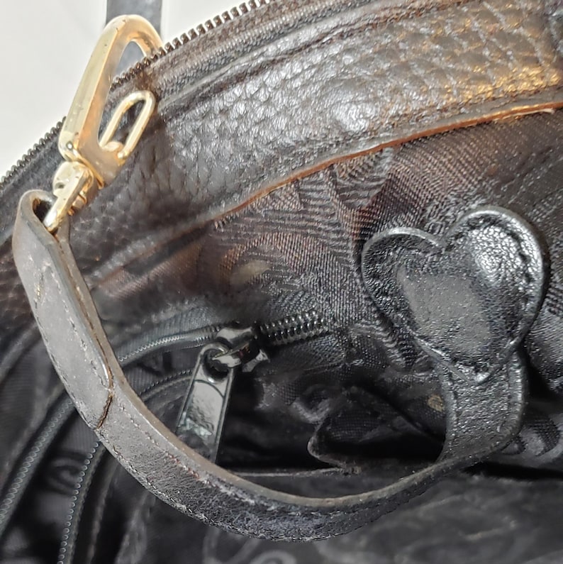 Puede incluir: Primer plano de un bolso de mano de cuero negro con un cierre y asa dorados. El bolso presenta un adorno en forma de coraz&oacute;n y un cierre de cremallera. El cuero tiene una apariencia texturizada.