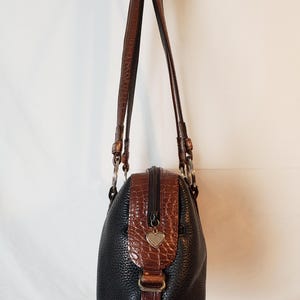 Puede incluir: Bolso de mano de cuero negro con detalles marrones con textura de cocodrilo. Cuenta con cremallera superior, correas de cuero marr&oacute;n y un dije en forma de coraz&oacute;n. El bolso tiene una forma estructurada y herrajes plateados.