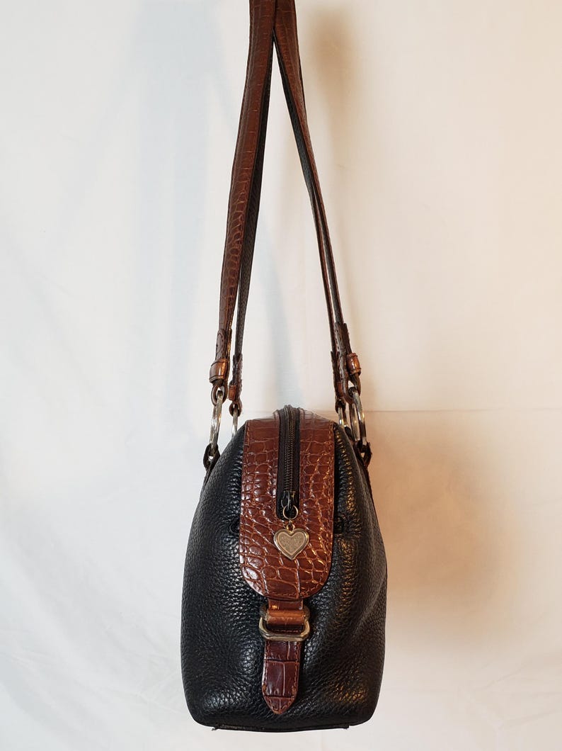 Puede incluir: Bolso de mano de cuero negro con detalles marrones con textura de cocodrilo. Cuenta con cremallera superior, correas de cuero marr&oacute;n y un dije en forma de coraz&oacute;n. El bolso tiene una forma estructurada y herrajes plateados.