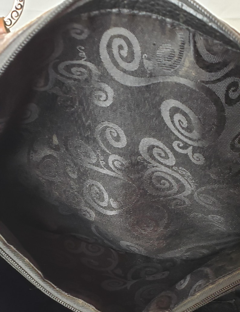 Puede incluir: Vista interior de un bolso marr&oacute;n con forro estampado. El forro presenta un dise&ntilde;o de remolino repetido en un tono m&aacute;s oscuro, creando un contraste sutil. El interior del bolso es visible, mostrando la textura y el dise&ntilde;o de la tela.