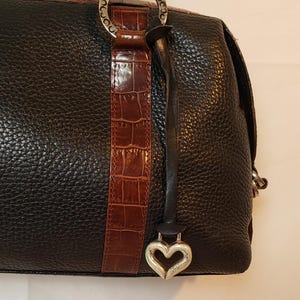 Puede incluir: Bolso de mano de cuero negro con acabado texturizado. Cuenta con una franja de cuero marr&oacute;n con relieve de cocodrilo y un dije plateado en forma de coraz&oacute;n. El bolso tiene herrajes plateados y un asa superior.