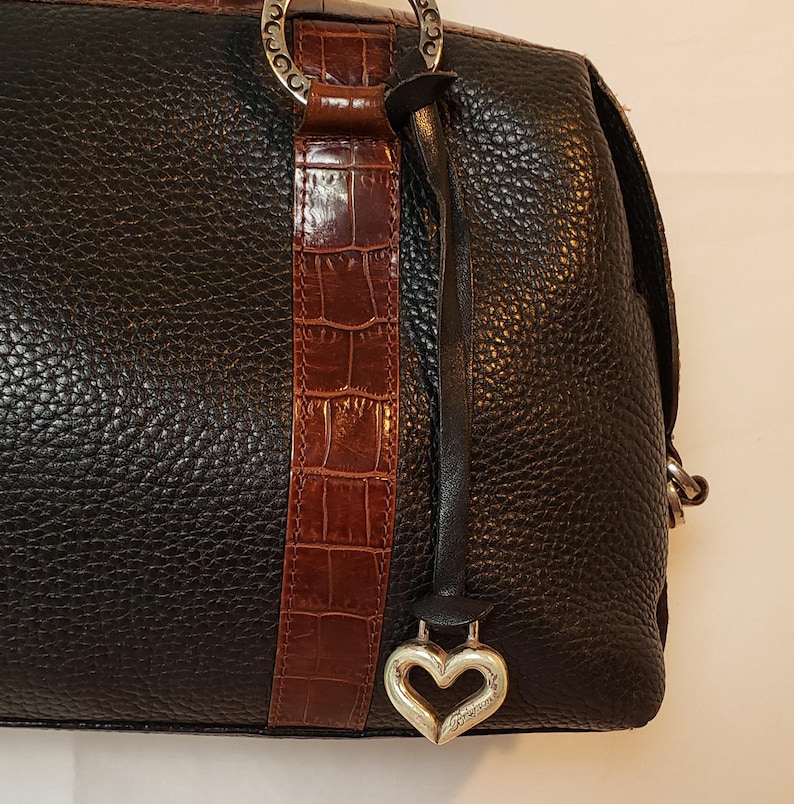 Puede incluir: Bolso de mano de cuero negro con acabado texturizado. Cuenta con una franja de cuero marr&oacute;n con relieve de cocodrilo y un dije plateado en forma de coraz&oacute;n. El bolso tiene herrajes plateados y un asa superior.
