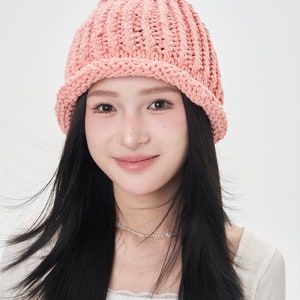 May include: A woman with long black hair is wearing a pink knitted beanie hat. The hat has a wide brim and a ribbed texture.