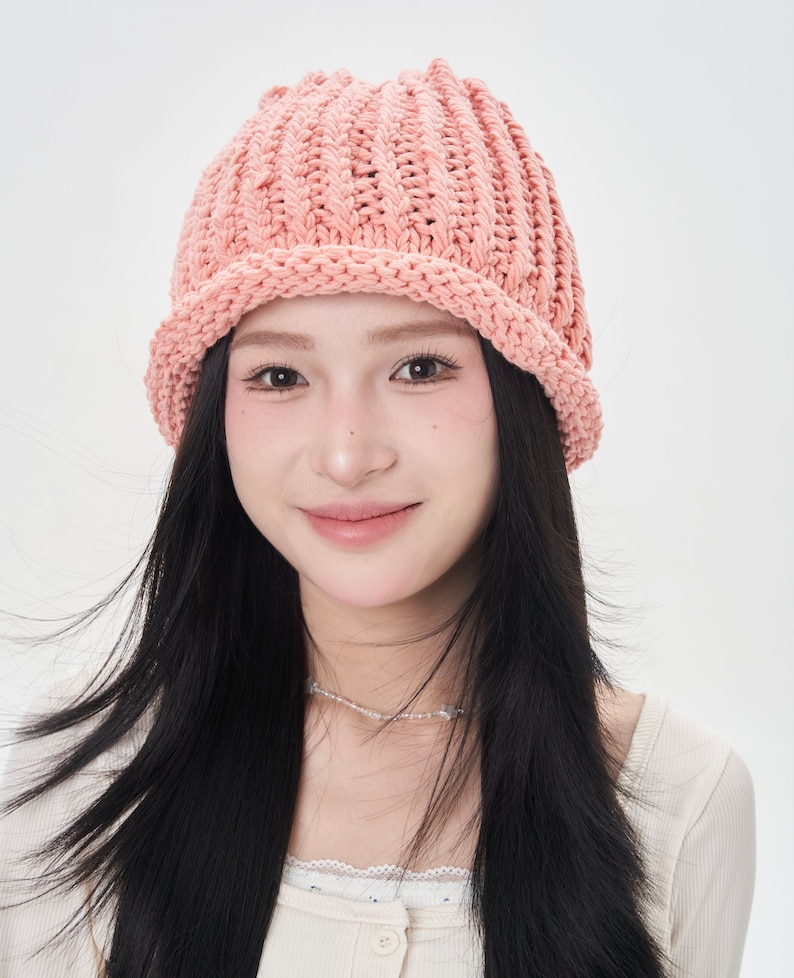 May include: A woman with long black hair is wearing a pink knitted beanie hat. The hat has a wide brim and a ribbed texture.