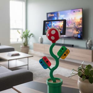 Puede incluir: Un soporte con forma de planta verde con una cabeza roja de puntos blancos que sostiene una consola de juegos. El tallo tiene dos ramas laterales, cada una con un mando colorido. La base es una maceta cilíndrica verde con una superficie plana beige.