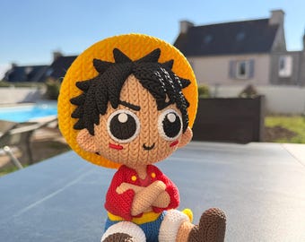 May include: A handmade Luffy doll, featuring a yellow straw hat, red shirt, blue shorts, and brown boots. The doll has black hair and large, expressive eyes. The doll is sitting on a dark surface, with a sunny outdoor background.