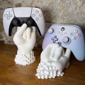 Peut inclure: Deux mains blanches imprimées en 3D tenant une manette PlayStation 5 blanche et une manette Xbox blanche. Les mains sont pixelisées et ont un design en blocs.
