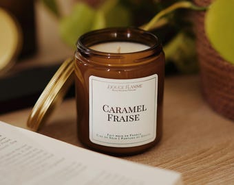 May include: A brown glass jar candle with a gold lid, labeled "CARAMEL FRAISE" with additional text. The candle sits next to an open book. The candle is made with soy wax and perfumes, and made in France.
