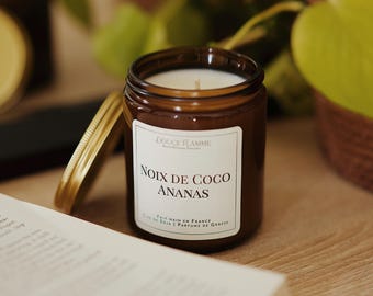 May include: A brown glass jar candle with a white label that reads "NOIX DE COCO ANANAS". The candle has a gold lid and a white wax interior. The text "DOUCE FLAMME" is at the top of the label. The candle is on a wooden surface.