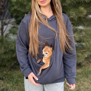 Puede incluir: Una sudadera con capucha gris oscuro con un bolsillo tipo canguro para llevar un perro pequeño. Un chihuahua marrón claro asoma por el bolsillo. La sudadera es de material suave, ideal para dueños de mascotas.