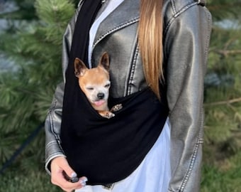 May include: A black pet carrier sling holds a small, light brown dog with its tongue sticking out. The dog carrier is worn over the shoulder. The person is wearing a silver leather jacket and a white shirt.