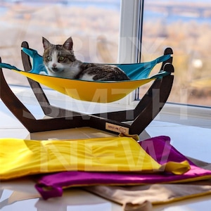 May include: A cat lounging in a yellow and blue hammock bed. The hammock is suspended from a wooden frame. The cat is looking at the camera. The hammock is on a white surface.