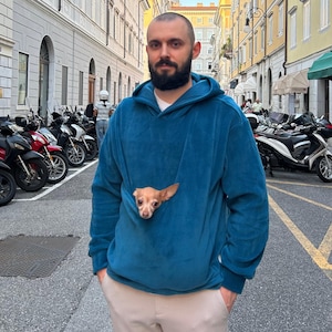 Könnte beinhalten: Ein tealblauer Kapuzenpullover mit einer Kängurutasche, in der ein kleiner hellbrauner Hund herausschaut. Der Kapuzenpullover wird von einer Person getragen, die auf einer Stadtstraße mit geparkten Motorrädern und Gebäuden im Hintergrund steht.