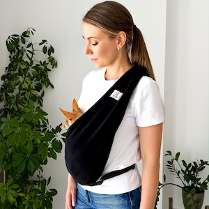 May include: A black fabric pet sling carrier worn across the chest, featuring a small white rectangular brand label. A small tan dog with pointed ears is visible inside the pouch. The carrier includes a black adjustable strap and buckle.