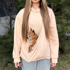 Op de afbeelding: Een lichtroze hoodie met een ingebouwde zak voor een kleine hond. Een kleine bruine en witte hond zit in de zak.