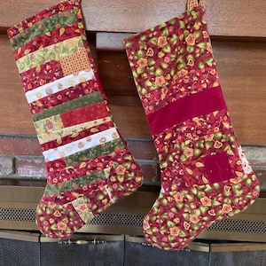 May include: Two Christmas stockings. One is patchwork with red, green, and white stripes. The other is red with a floral pattern and a burgundy velvet band. Both have fabric loops for hanging.