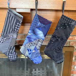 May include: Three Christmas stockings constructed from repurposed neckties in various shades of blue, gray, and black. Each stocking features a patchwork design of geometric, striped, and polka dot patterns, with a fabric loop at the top for hanging.