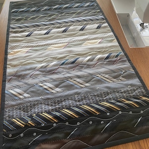May include: A table runner made from a variety of patterned ties. The runner is made in a gradient pattern, starting with dark blue and gray ties and transitioning to lighter colors, ending with black and gold ties. The runner is edged with a black fabric.