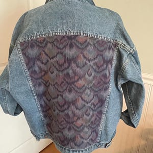 May include: A light blue denim jacket with a patterned fabric panel on the back. The panel features a design of purple, blue, and brown swirls.