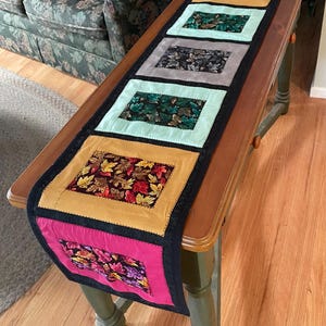 May include: A table runner with a patchwork design featuring six rectangular panels. Each panel is a different color and fabric, with a floral pattern. The panels are sewn together with black trim. The table runner is on a wooden table.