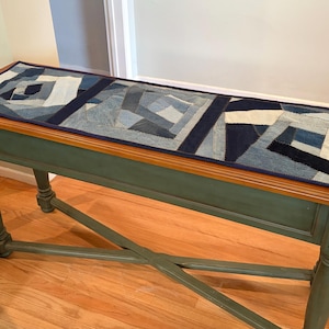 May include: A rectangular denim patchwork table runner with a dark blue border rests on a green and brown wooden table. The runner features geometric patterns in various shades of blue and white. The table has turned legs and an X-shaped support.