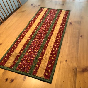 May include: A table runner with a striped pattern in shades of red, green, and beige. The runner is made of fabric with a floral print and has a green border.