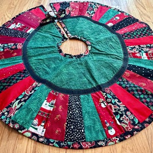 May include: Circular Christmas tree skirt with a green center and a black border. The skirt features alternating panels of red, green, and black fabric with festive patterns, including snowmen and floral designs.