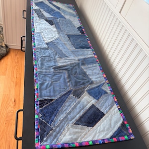 May include: A rectangular table runner constructed from various shades of blue denim fabric scraps in a patchwork design. The runner features visible stitching patterns and is finished with a colorful, multi-patterned fabric border trim.