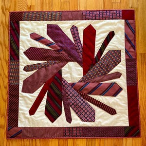 May include: A patchwork quilt with a central design of a flower made from various shades of red and burgundy neckties. The quilt is framed with a border of the same neckties.