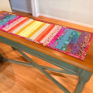 May include: A rectangular quilted table runner featuring a vibrant spectrum of vertical fabric strips in shades of yellow, orange, pink, purple, and blue. The runner has a thin red border and a multi-colored patterned backing visible at the edges.