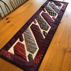 May include: A table runner made from repurposed neckties. The runner is made of various patterned ties in shades of red, blue, and white. The runner is trimmed with a black fabric border.