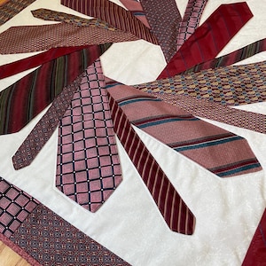 May include: A white quilt with a design of red, burgundy, and pink ties arranged in a circular pattern. The ties are sewn together to create a unique and decorative design.