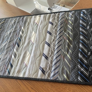 May include: A table runner made from repurposed ties in shades of gray, black, and white. The ties are sewn together in a vertical pattern and quilted with a wavy design.