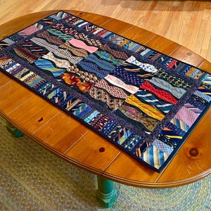 May include: A table runner made from a patchwork of colorful neckties. The neckties are sewn together in a wavy pattern and are arranged in a variety of colors and patterns. The runner is on a wooden table.