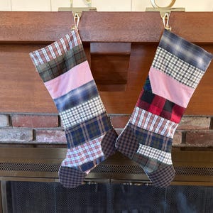 May include: Two Christmas stockings hanging from a wooden mantel. Each stocking is made of patchwork fabric in various plaid and patterned designs, including pink, red, and blue. The stockings are suspended by gold-colored metal hooks.