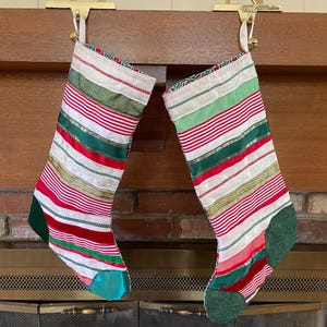 May include: Two Christmas stockings with red and white stripes, green and gold ribbons, and green heels and toes hang from a wooden mantel. The stockings are suspended by gold-colored metal stocking holders.