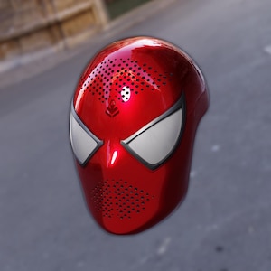 May include: A glossy red mask with white eye shapes and a pattern of small black dots. The mask has a smooth, reflective surface and a design reminiscent of a superhero. The mask is likely a costume accessory.