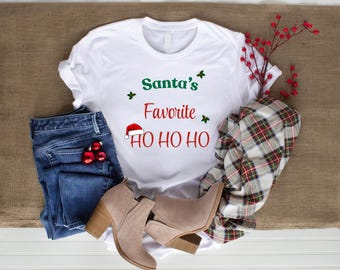 May include: White t-shirt with the text "Santa's Favorite HO HO HO" in green and red. The shirt is paired with blue jeans, tan ankle boots, and a red and green plaid scarf. Christmas-themed apparel.