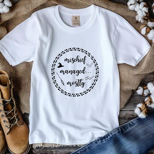 May include: White t-shirt with the text "mischief managed... mostly" in a black circular design. The design includes a witch hat, a broom, glasses, and wings. The shirt is displayed on a rustic surface with boots and denim.