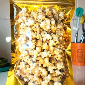 May include: A clear, rectangular stand-up pouch with a shiny gold metallic border filled with caramel-coated popcorn. The bag features a resealable zipper top and a small circular hole at the top center for hanging, showcasing the golden-brown snack.